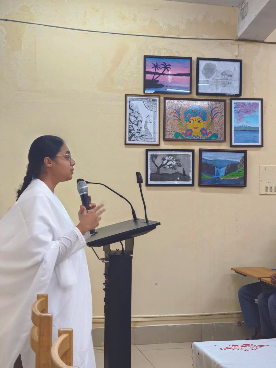 Smriti Budhia speaking at a podium, with student artwork displayed on the wall behind her
